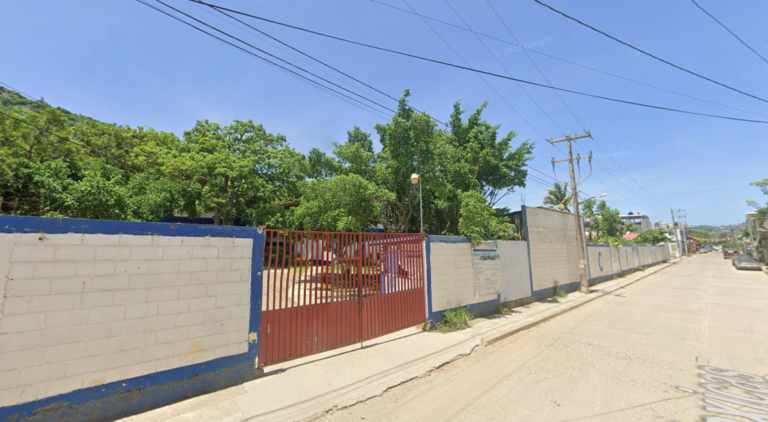Fotografía en la que se muestra la entrada del plantel, conformada por muros de color blanco con bordes azules que rodean las instalaciones. En medio de dos muros hay una reja de color rojo por la que se puede ingresar al plantel.